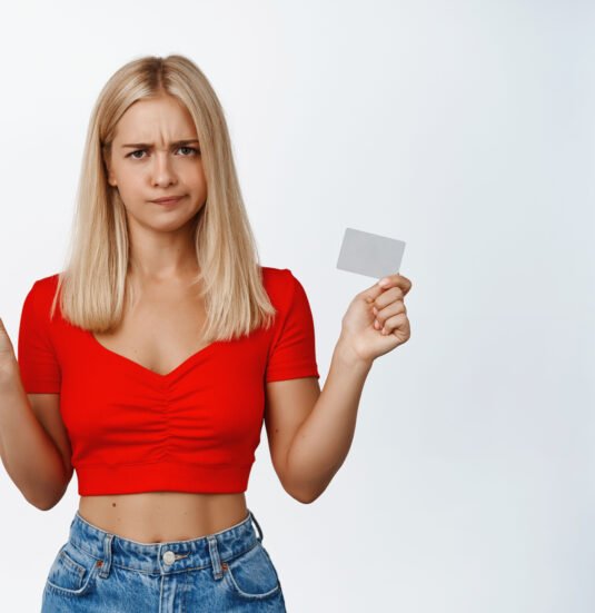 Displeased blond girl grimaces, shows mobile phone screen and credit card, pouting upset, standing over white background